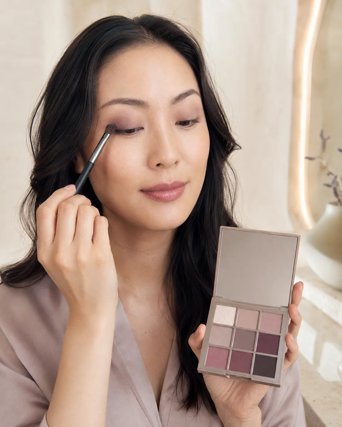 Woman applying mauve eyeshadow while holding an unbranded nine-pan palette